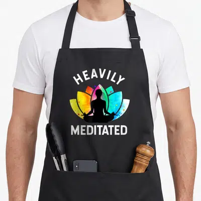 Heavily stepping into apron - Black meditating silhouette over yellow red blue watercolor lotus with Heavily Meditated — shown on a black cotton canvas apron against a white background