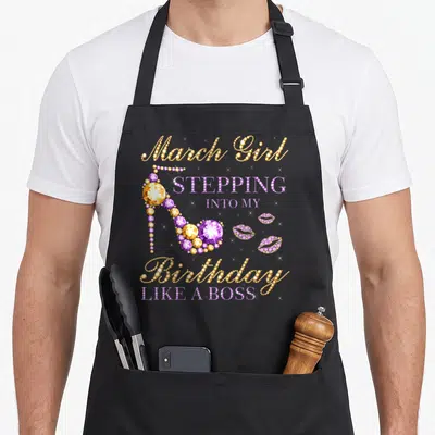 March bbq boss apron - Rhinestone text March Girl Stepping Into My Birthday Like A Boss with purple gold high heel — shown on a black cotton canvas apron against a white background