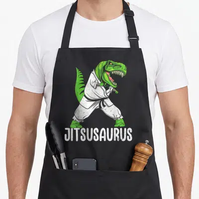 Jitsusaurus nourjoy apron - Green dinosaur in white gi and black belt with JITSUSAURUS text — shown on a black cotton canvas apron against a white background