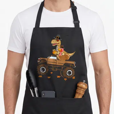 Dinosaur thanksgiving apron - T-Rex and turkey in pilgrim hats riding a brown monster truck with scattered orange leaves — shown on a black cotton canvas apron against a white background
