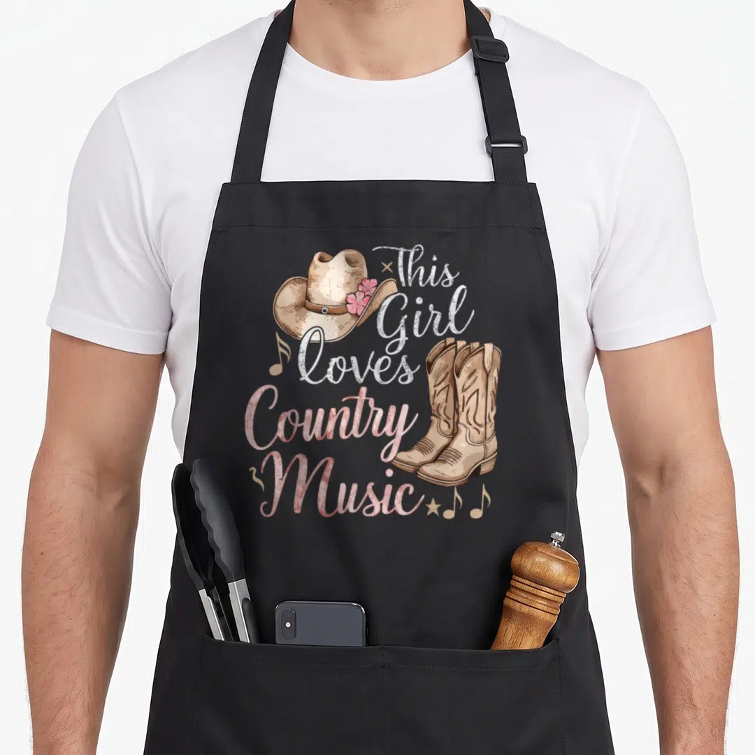 Retro music cook's country aprons - Stacked text This Girl Loves Country Music with cowboy hat, boots, and musical notes — shown on a black cotton canvas apron against a white background