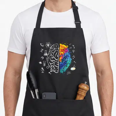 Brain science apron - Split brain with white science icons on left and multicolored art splash on right — shown on a black cotton canvas apron against a white background