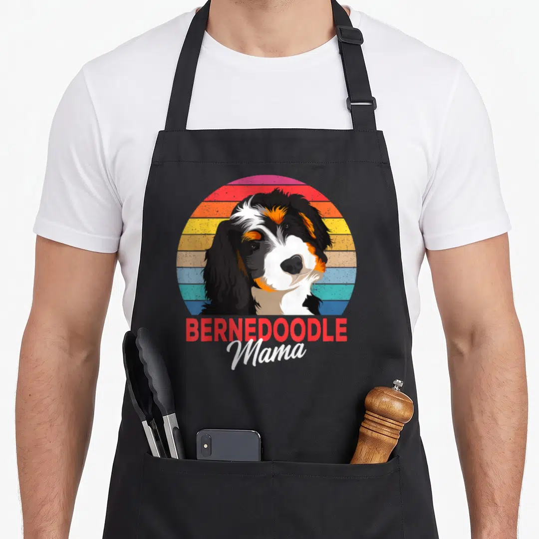 Bernedoodle mama dog apron - Cartoon Bernedoodle head inside a red orange and blue striped circle with text below — shown on a black cotton canvas apron against a white background