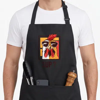 Gardening chicken coop apron - Cartoon rooster with sunglasses in brown frame above text SUP ? in red and orange — shown on a black cotton canvas apron against a white background