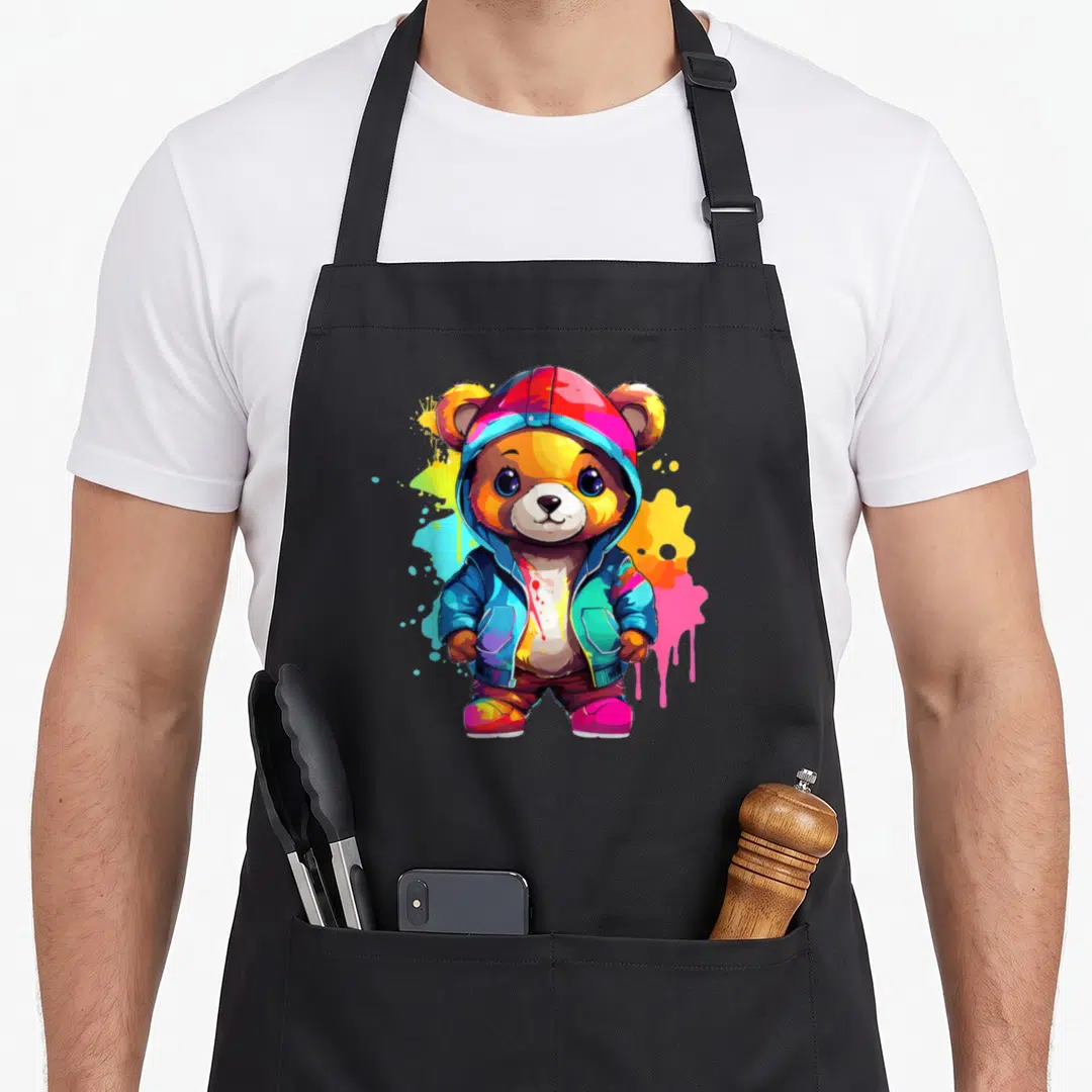 Colorful teddy bear apron - Cartoon bear in hoodie surrounded by blue, orange, pink, yellow paint splatters — shown on a black cotton canvas apron against a white background