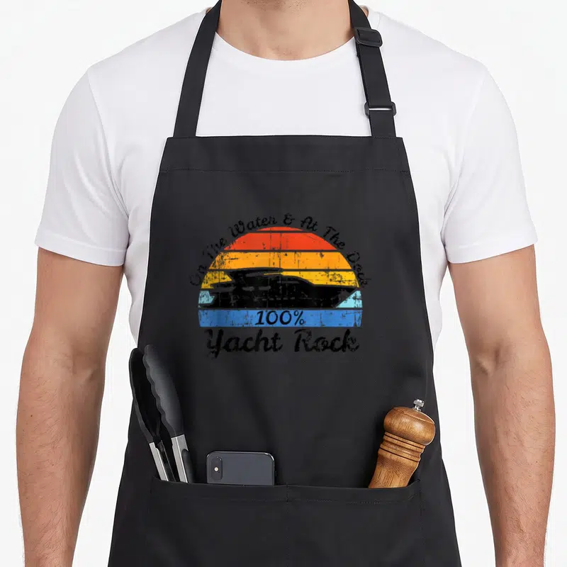 Yacht rock distressed apron - Retro striped sunset with black yacht silhouette and Yacht Rock text — shown on a black cotton canvas apron against a white background