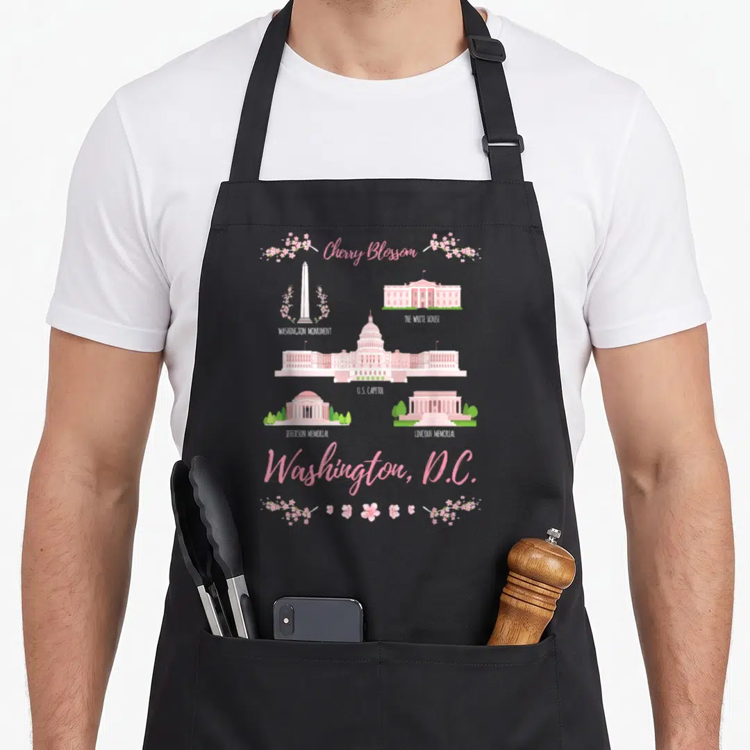 Monument cherry apron - Pink cartoon Washington DC landmarks with cherry blossom branches and text labels — shown on a black cotton canvas apron against a white background