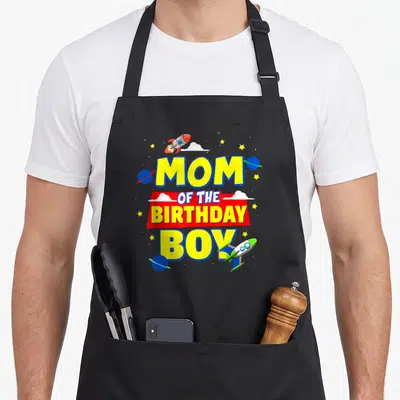Astronaut outer birthday apron - MOM OF THE BIRTHDAY BOY text with rockets, planets, stars and clouds in yellow red — shown on a black cotton canvas apron against a white background