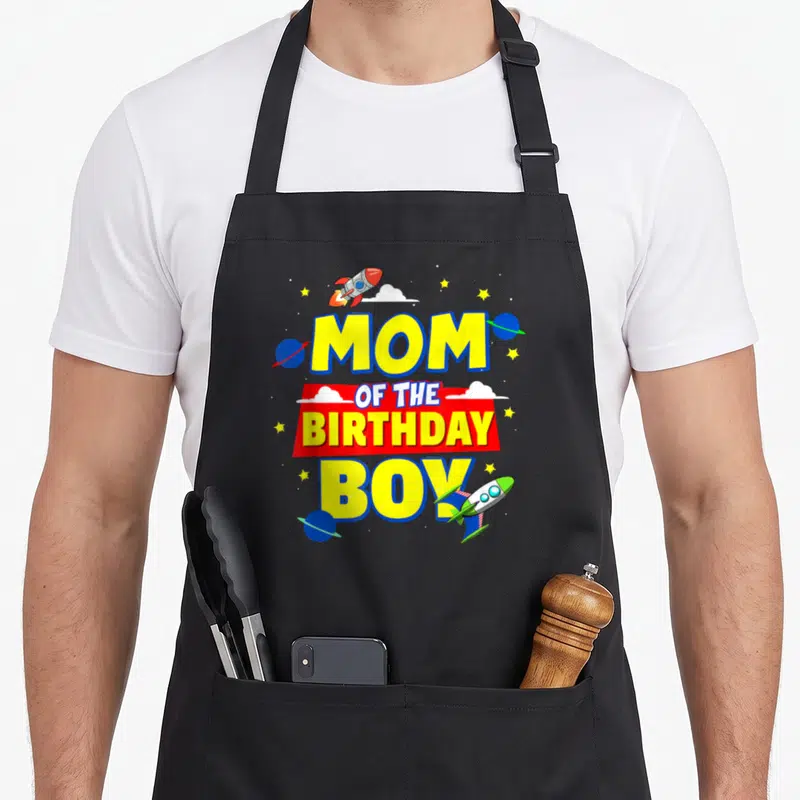 Astronaut outer birthday apron - MOM OF THE BIRTHDAY BOY text with rockets, planets, stars and clouds in yellow red — shown on a black cotton canvas apron against a white background