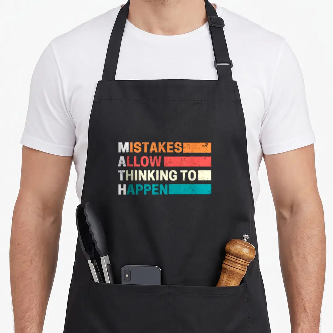 Math algebra teacher apron - Stacked white text MISTAKES ALLOW THINKING TO HAPPEN overlapped by distressed orange, red — shown on a black cotton canvas apron against a white background