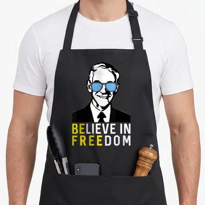 Believe liberty apron - Stylized man in blue glasses above BELIEVE IN FREEDOM text in yellow and white — shown on a black cotton canvas apron against a white background