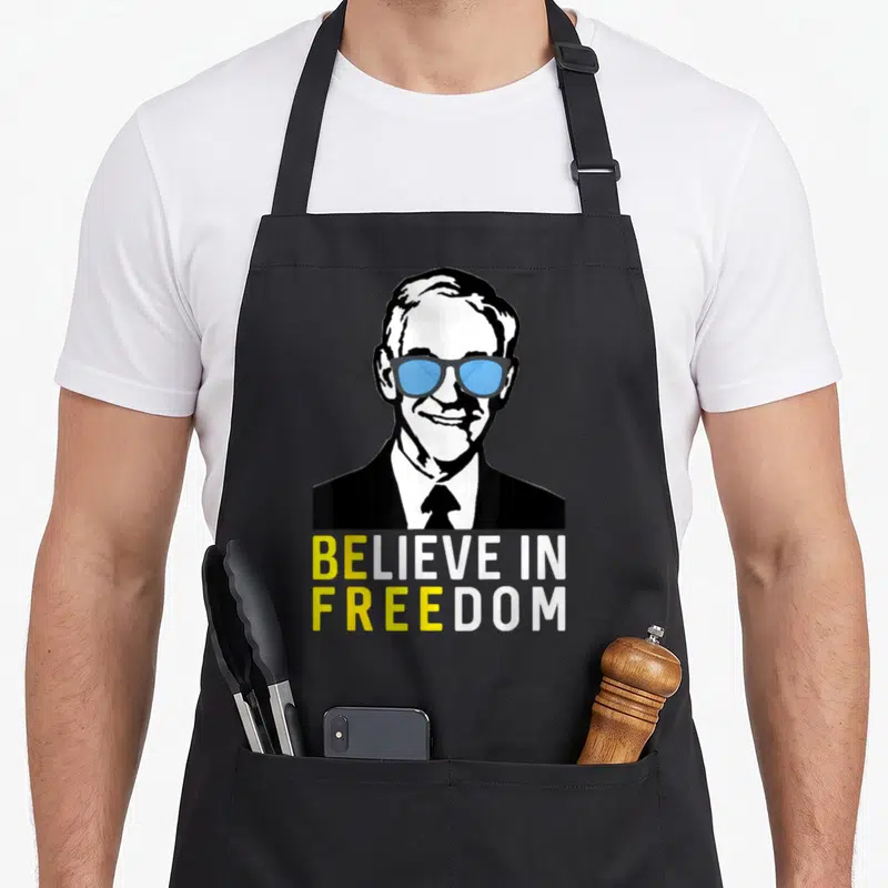 Believe liberty apron - Stylized man in blue glasses above BELIEVE IN FREEDOM text in yellow and white — shown on a black cotton canvas apron against a white background