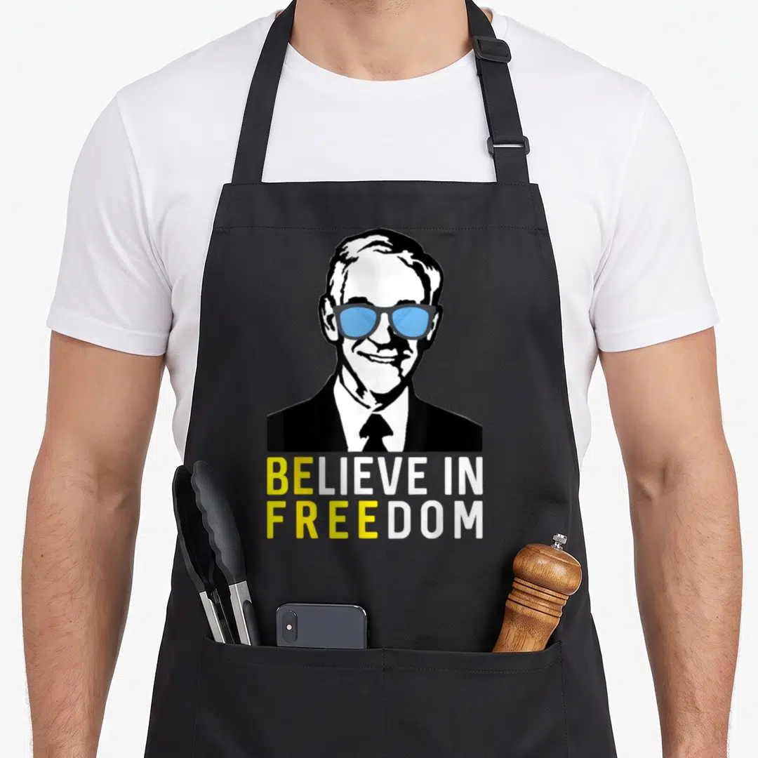 Believe liberty apron - Stylized man in blue glasses above BELIEVE IN FREEDOM text in yellow and white — shown on a black cotton canvas apron against a white background