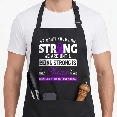 Warrior domestic aprons - Vertical white text with purple ribbon replacing O and purple banner at bottom — shown on a black cotton canvas apron against a white background
