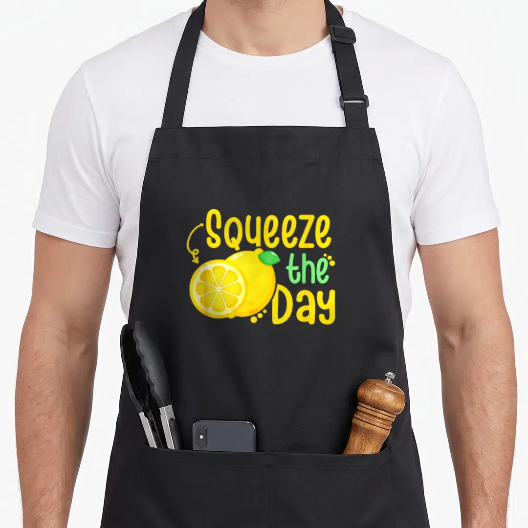 Summer lemon apron - Squeeze the Day text with whole and sliced yellow lemon and green leaf — shown on a black cotton canvas apron against a white background