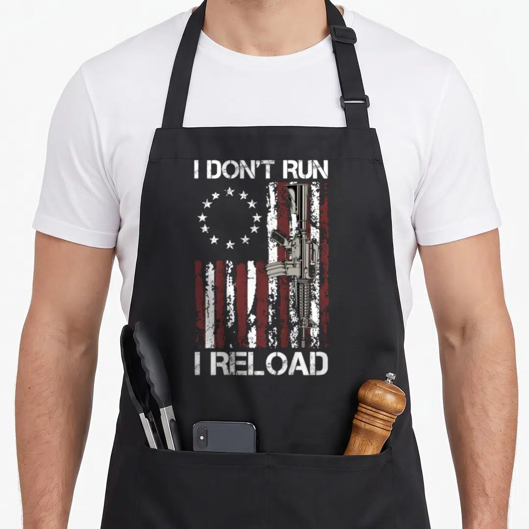Run gun apron - I don't run I reload text over distressed flag and rifle — shown on a black cotton canvas apron against a white background