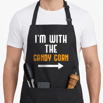 Candy corn costume halloween apron - Stacked white and orange text I'M WITH THE CANDY CORN above a white arrow pointing — shown on a black cotton canvas apron against a white background