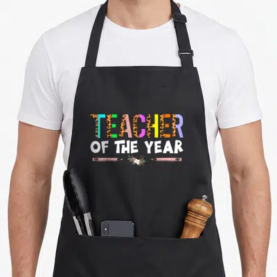 Cute appreciation teacher apron - Multicolored TEACHER OF THE YEAR text with pink pencils and floral accent — shown on a black cotton canvas apron against a white background