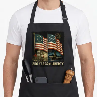 Betsy birthday apron - Two USA flags, sailing ship, aircraft carrier, 250 YEARS OF LIBERTY text, gold fireworks — shown on a black cotton canvas apron against a white background