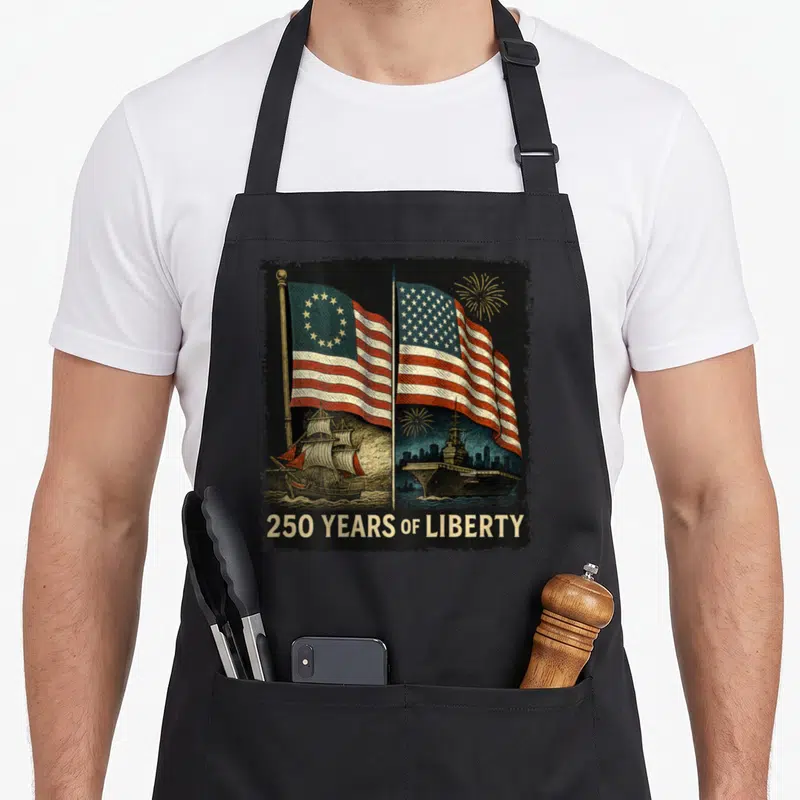 Betsy birthday apron - Two USA flags, sailing ship, aircraft carrier, 250 YEARS OF LIBERTY text, gold fireworks — shown on a black cotton canvas apron against a white background