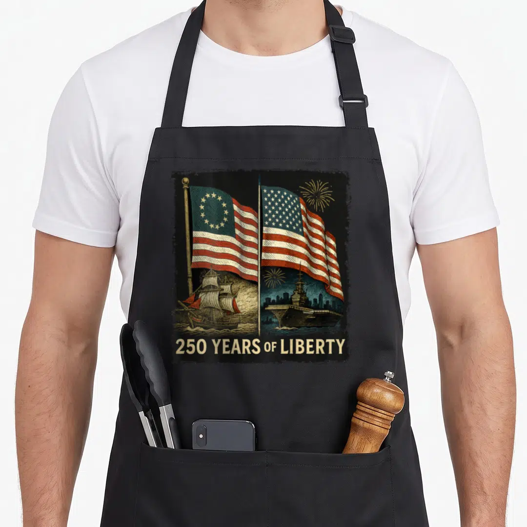 Betsy birthday apron - Two USA flags, sailing ship, aircraft carrier, 250 YEARS OF LIBERTY text, gold fireworks — shown on a black cotton canvas apron against a white background