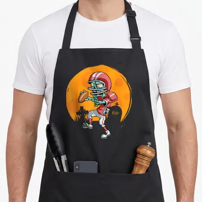 Football halloween apron - Cartoon skeleton football player holding a ball near tombstones with RIP text on orange — shown on a black cotton canvas apron against a white background