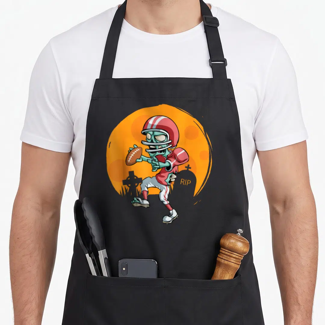 Football halloween apron - Cartoon skeleton football player holding a ball near tombstones with RIP text on orange — shown on a black cotton canvas apron against a white background
