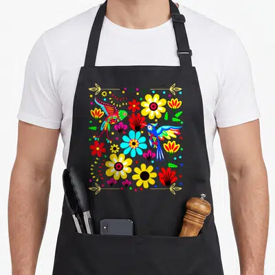 Bird the floral apron - Two cartoon hummingbirds facing each other surrounded by yellow, red, blue, and green flowers — shown on a black cotton canvas apron against a white background