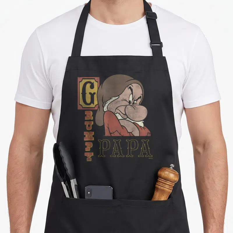 Snow papa apron - Grumpy dwarf illustration with vertical GRUMPY text and horizontal PAPA label in distressed style — shown on a black cotton canvas apron against a white background