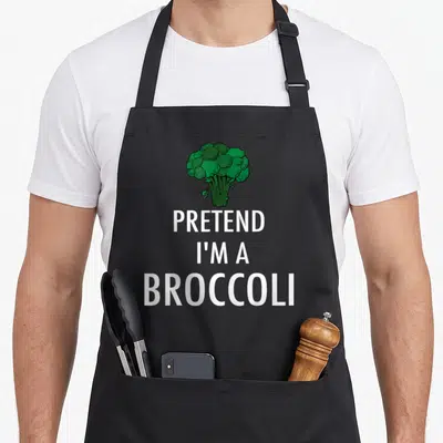 Pretend broccoli halloween apron - Cartoon green broccoli centered above stacked white text reading PRETEND I'M — shown on a black cotton canvas apron against a white background