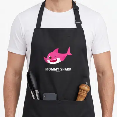 Shark pinkfong apron - Pink cartoon shark winking above white MOMMY SHARK text on black fabric — shown on a black cotton canvas apron against a white background