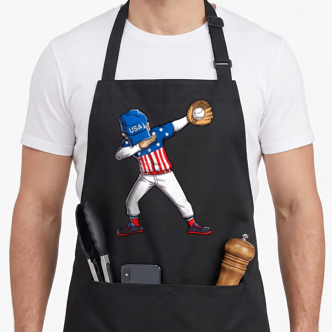 Player 4th of july apron - Cartoon baseball player dabbing in USA uniform holding glove with ball — shown on a black cotton canvas apron against a white background
