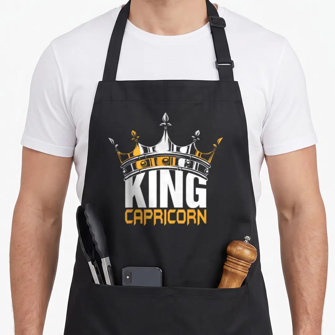 Capricorn king apron - Cartoon crown above white KING and orange CAPRICORN text — shown on a black cotton canvas apron against a white background