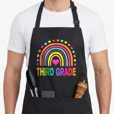 Grade 3rd art teacher aprons - Rainbow arch with pink heart and THIRD GRADE text in orange pink purple yellow blue green — shown on a black cotton canvas apron against a white background