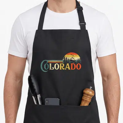 Sun colorado apron - Retro sun and mountain silhouette overlapping multicolored COLORADO text in teal and orange — shown on a black cotton canvas apron against a white background
