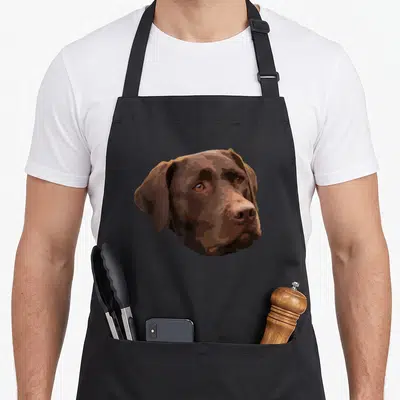 Lab retriever chocolate labrador apron - Cartoon brown dog head illustration centered on canvas apron — shown on a black cotton canvas apron against a white background