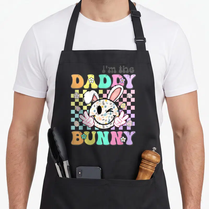 Groovy easter bunny aprons - i'm the DADDY BUNNY text with patterned smiley face, peace hands, and checkerboard — shown on a black cotton canvas apron against a white background