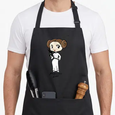 Funny portrait cartoon apron - Cartoon Princess Leia in white dress holding black blaster with text below — shown on a black cotton canvas apron against a white background