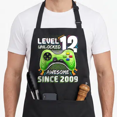 Unlocked 12th video game apron - Green game controller with Level 12 Unlocked and Awesome Since 2009 text — shown on a black cotton canvas apron against a white background