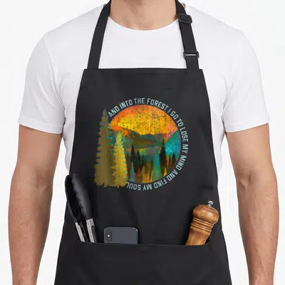 Nature camping apron - Retro forest landscape with circular text arch AND INTO THE FOREST I GO TO LOSE MY MIND AND FIND — shown on a black cotton canvas apron against a white background