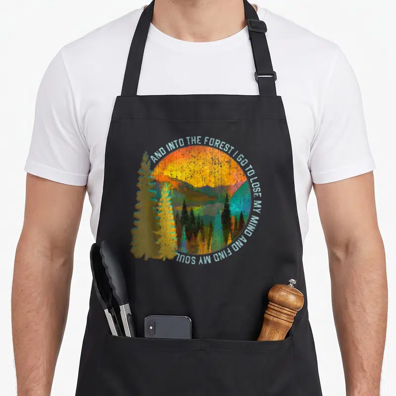Nature camping apron - Retro forest landscape with circular text arch AND INTO THE FOREST I GO TO LOSE MY MIND AND FIND — shown on a black cotton canvas apron against a white background