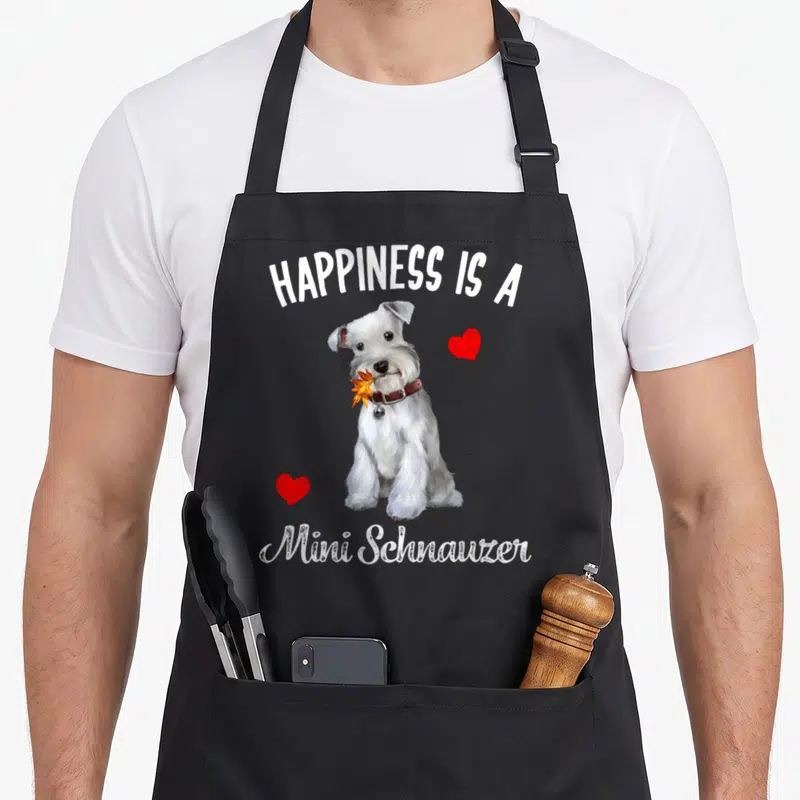 Cute dog miniature schnauzer apron - Realistic white Miniature Schnauzer holding an orange leaf between two red hearts — shown on a black cotton canvas apron against a white background