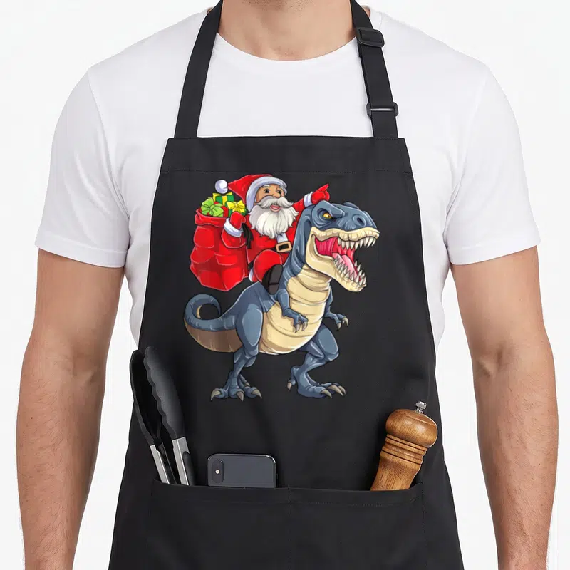 Dinosaur christmas santa apron - Santa Claus riding a blue T-Rex holding a red sack of gifts in a cartoon style — shown on a black cotton canvas apron against a white background