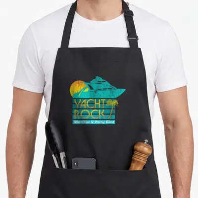 Rock yacht apron - Blue yacht silhouette over yellow sun with Yacht Rock text and palm tree — shown on a black cotton canvas apron against a white background