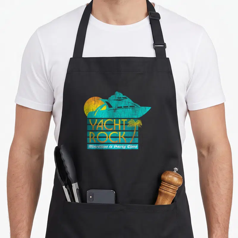 Rock yacht apron - Blue yacht silhouette over yellow sun with Yacht Rock text and palm tree — shown on a black cotton canvas apron against a white background