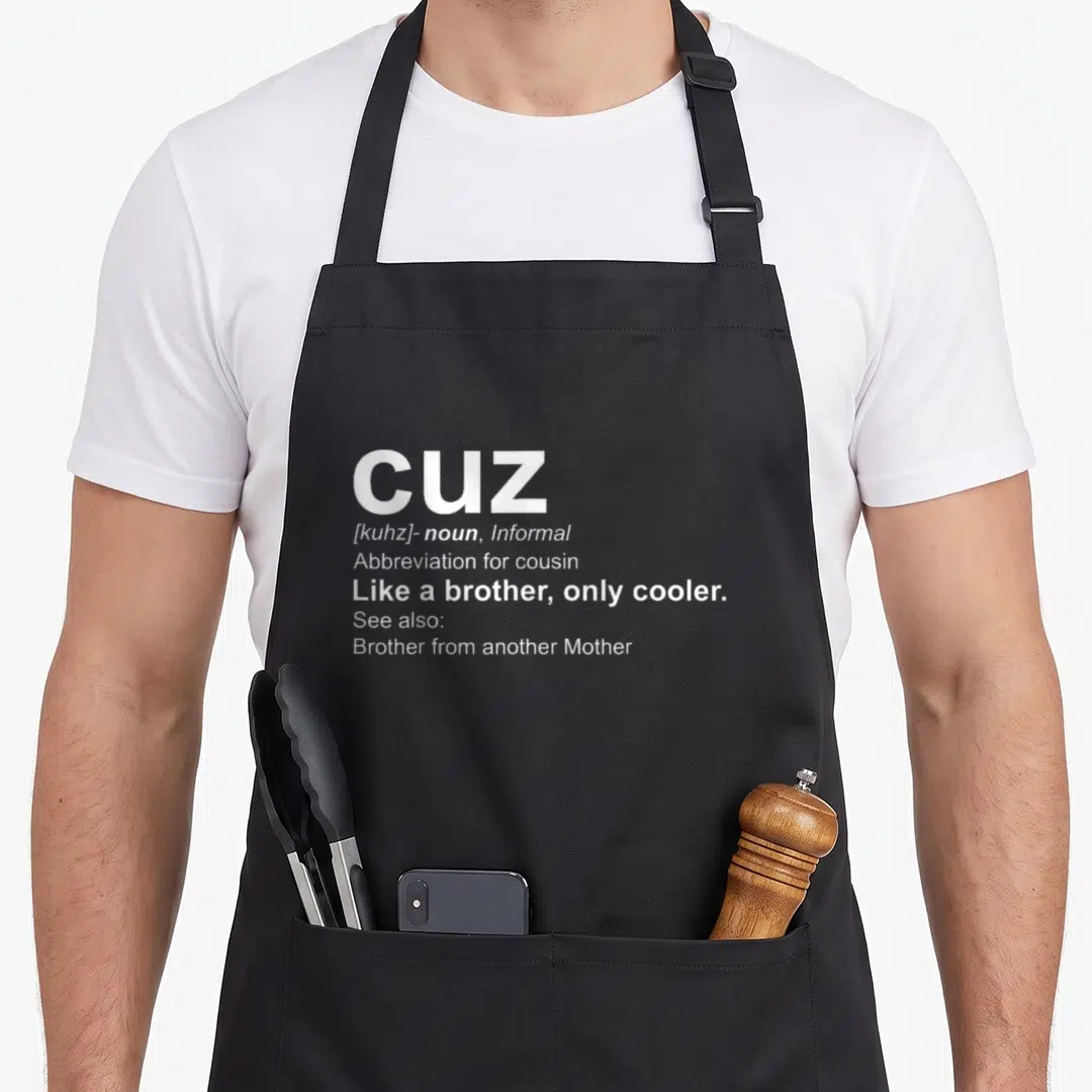 Cuz cousin apron - White dictionary style typography defining CUZ as like a brother only cooler — shown on a black cotton canvas apron against a white background