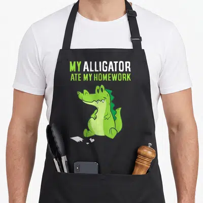 Crocodile ate homework apron - Green cartoon alligator with white paper scraps and stacked text — shown on a black cotton canvas apron against a white background