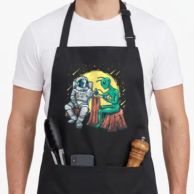 Astronaut tattoo apron - Green alien tattooing white astronaut on brown stumps under yellow moon — shown on a black cotton canvas apron against a white background