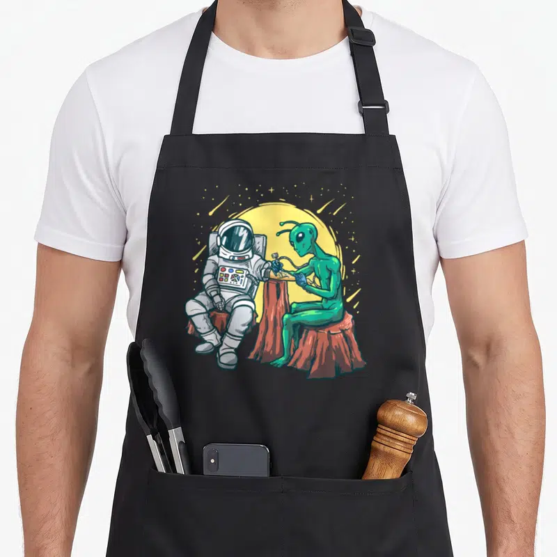Astronaut tattoo apron - Green alien tattooing white astronaut on brown stumps under yellow moon — shown on a black cotton canvas apron against a white background