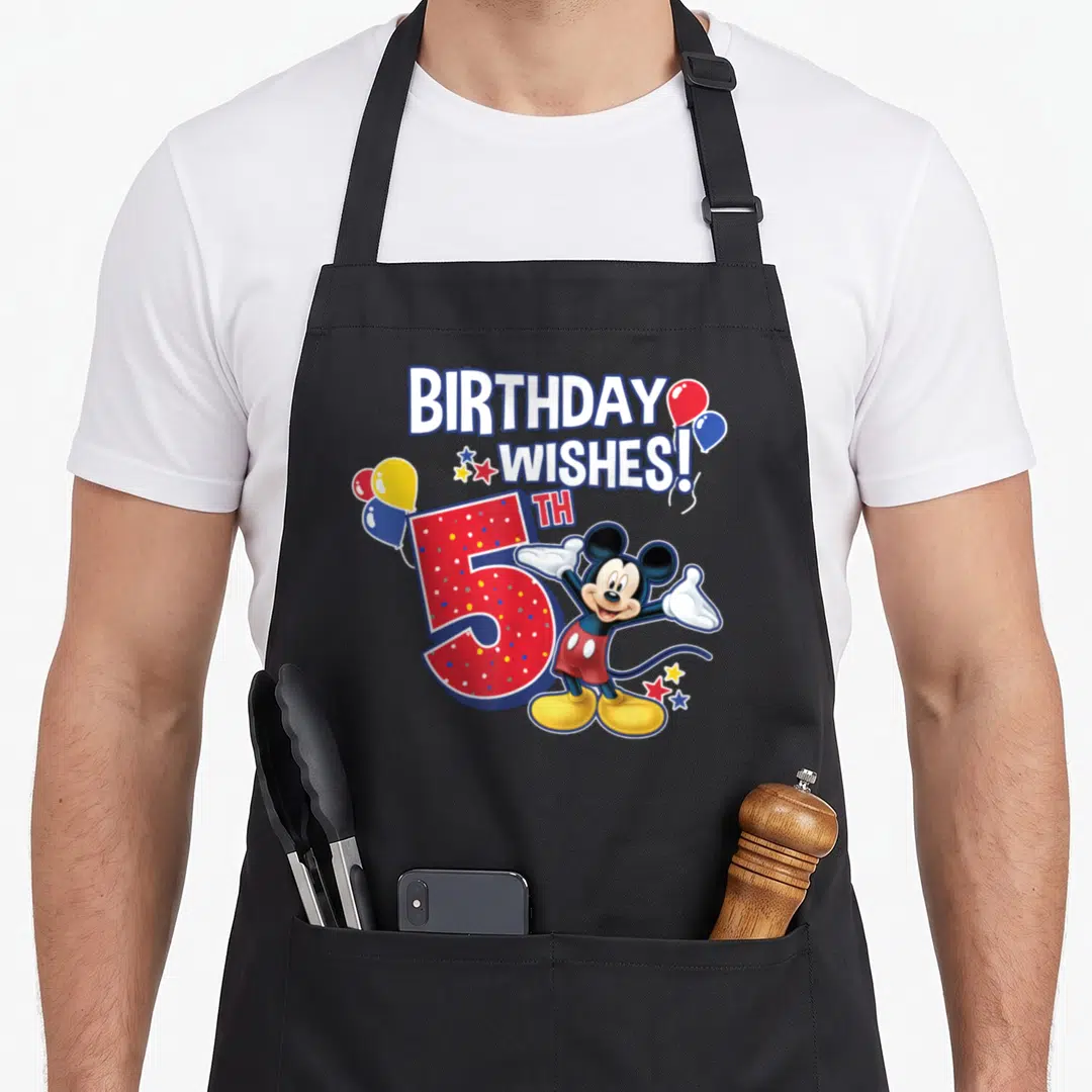 5th friends apron - Mickey Mouse stands next to a red number 5 and BIRTHDAY WISHES text with balloons and stars — shown on a black cotton canvas apron against a white background